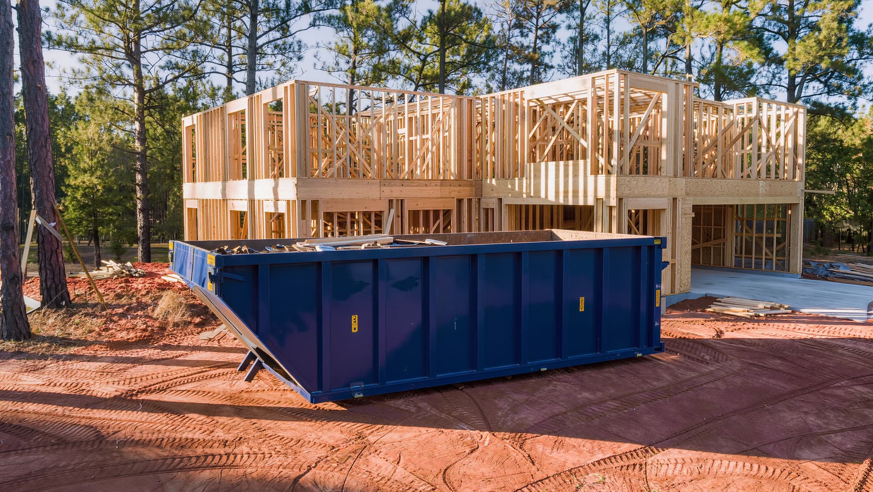 Large roll-off dumpster with scrap lumber beside a framed house on red clay